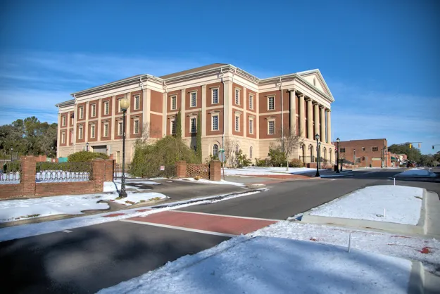 Large brick neoclassical building with tall columns and green landscaping on a snowy winter day under a clear blue sky